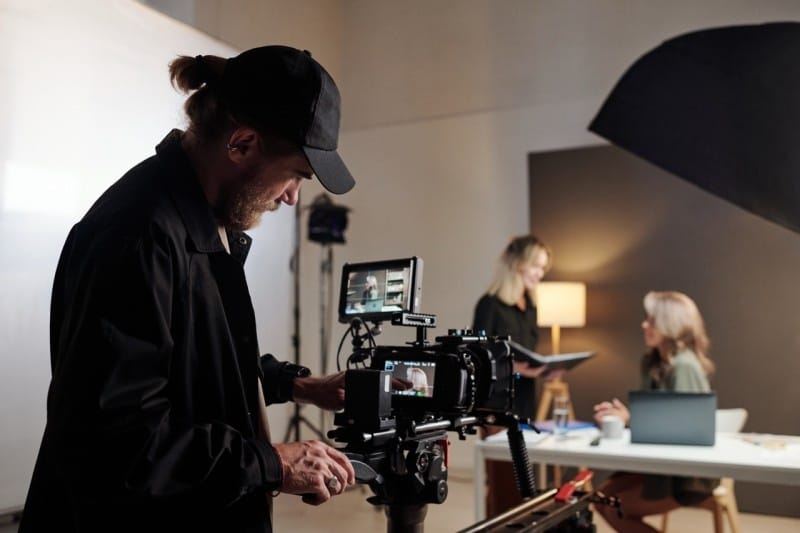 A film crew member operates a camera while two women engage in discussion during a shoot.