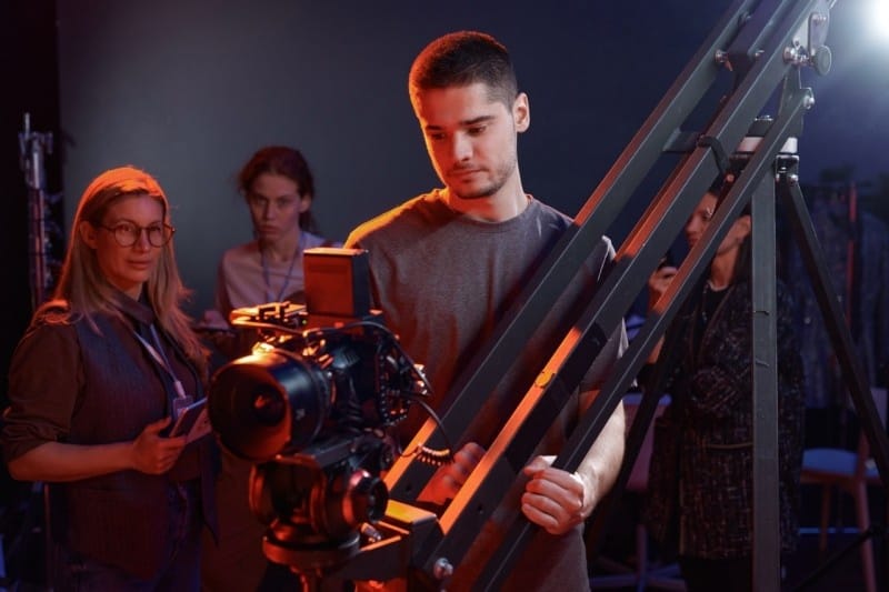 A photography setup showing a man adjusting a camera on a crane while two women observe in a studio environment.