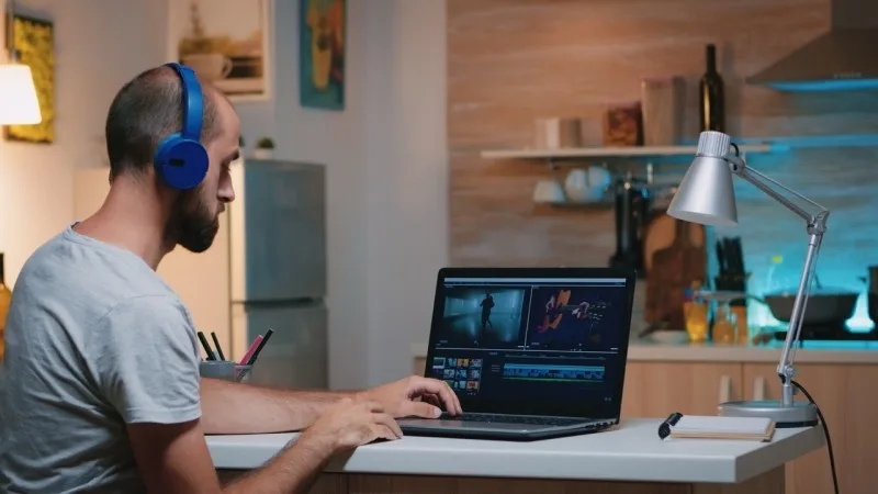 A person editing videos on a laptop in a modern kitchen environment with blue headphones on and a focused expression