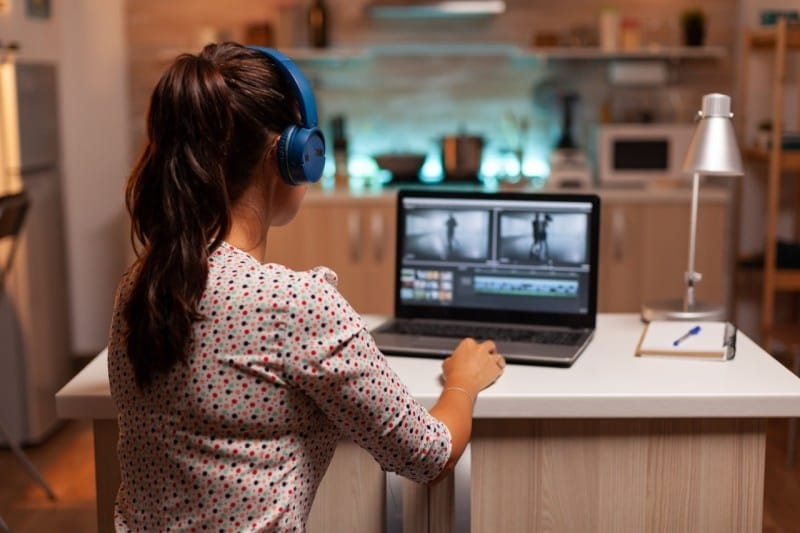 A person wearing blue headphones is editing a video on a laptop in a cozy kitchen setting