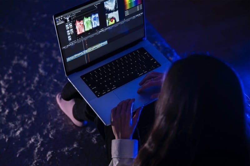 A person using a laptop to edit video footage in a dimly lit room, highlighting video editing software tools.