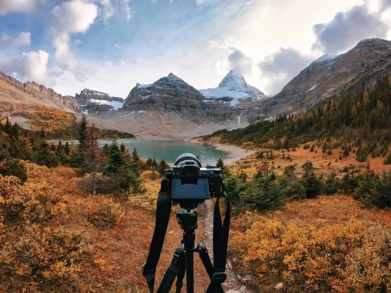 A camera set on a tripod captures a stunning autumn landscape with mountains, a lake, and colorful foliage.