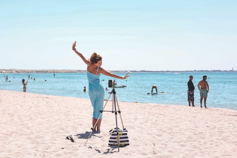 A woman in a blue dress poses elegantly on a beach while taking photos with a camera on a tripod, surrounded by people enjoying the sun and water.