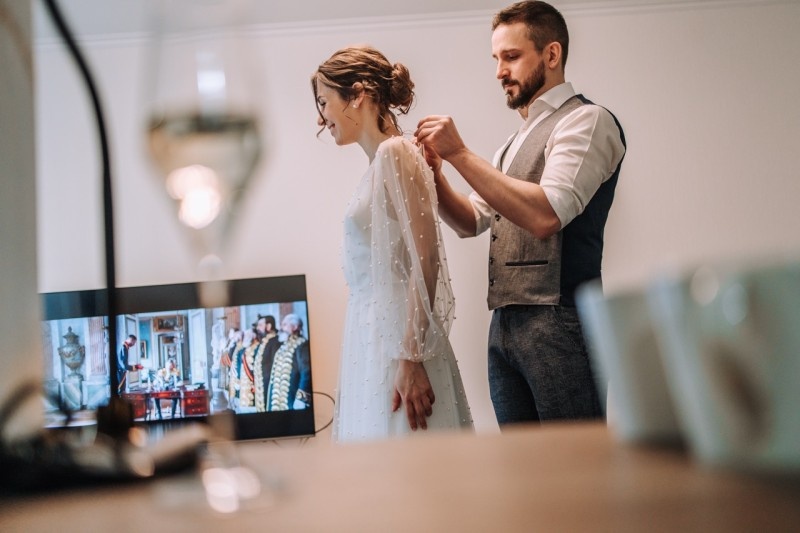 A couple preparing for a special occasion, as the man helps the woman with her dress while a movie plays in the background.