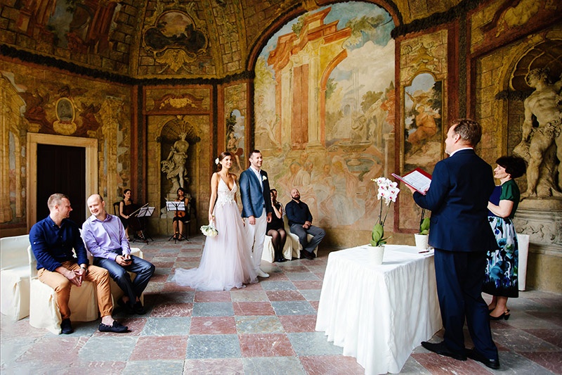 A beautiful wedding ceremony taking place in a historic venue with intricate wall paintings, featuring a happy couple standing before officiants and guests.