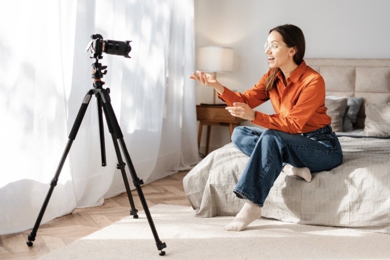 A woman in an orange shirt engaged in a live streaming session, animatedly talking to the camera on a tripod in a cozy bedroom setting.