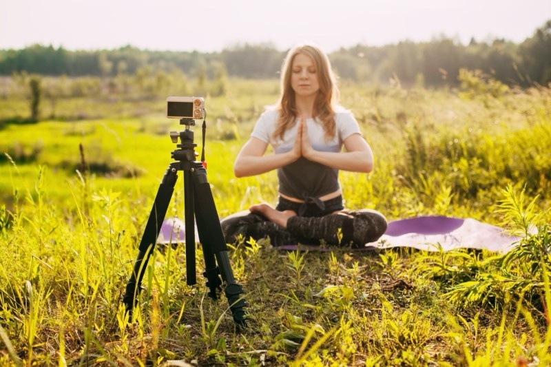 A woman meditating outdoors in a peaceful meadow, practicing mindfulness and yoga with a camera on a tripod.