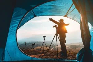 Best Travel Tripods 2025: Top Picks & Tips for Photo & Video. A photographer enjoying nature at sunrise, framed by a tent, with several tripods and cameras set up outside.