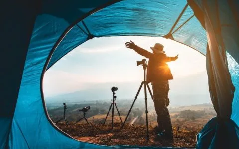 Best Travel Tripods 2025: Top Picks & Tips for Photo & Video. A photographer enjoying nature at sunrise, framed by a tent, with several tripods and cameras set up outside.