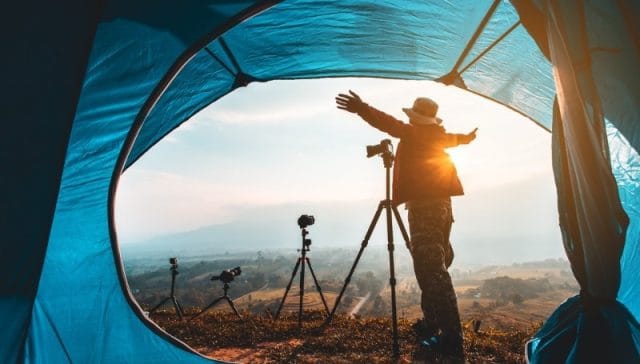 Best Travel Tripods 2025: Top Picks & Tips for Photo & Video. A photographer enjoying nature at sunrise, framed by a tent, with several tripods and cameras set up outside.