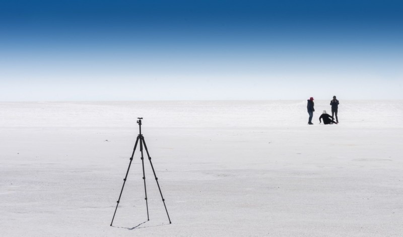 A camera tripod positioned on a stark white salt flat with people capturing photos in the background.