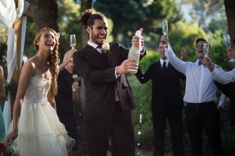 A joyful couple celebrating their wedding, with the groom joyfully popping a champagne bottle and guests raising their glasses in a beautiful outdoor setting.