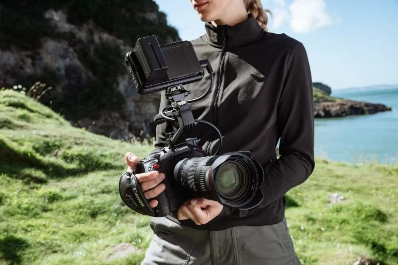 A filmmaker holding a Canon camera with a large lens, preparing to shoot in a scenic outdoor location by the water.