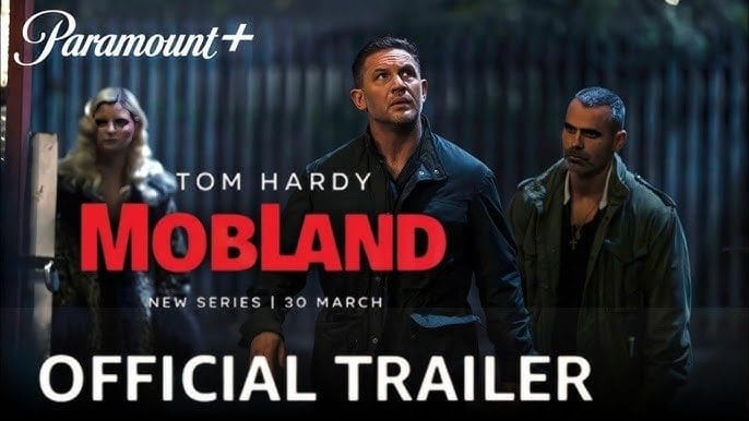 A scene from the official trailer of 'Mobland' featuring Tom Hardy with a dramatic expression, surrounded by characters, set in a suspenseful atmosphere.